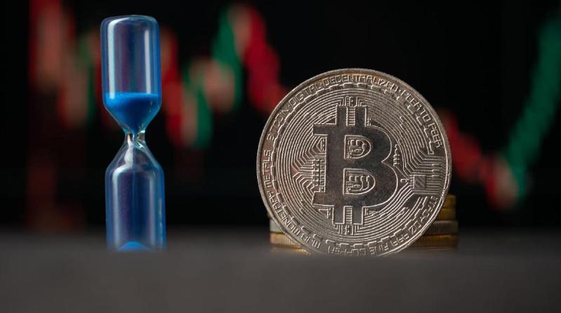 An image of a bitcoin (BTC) coin with an hourglass next to it and a graph of a volatile price in the background